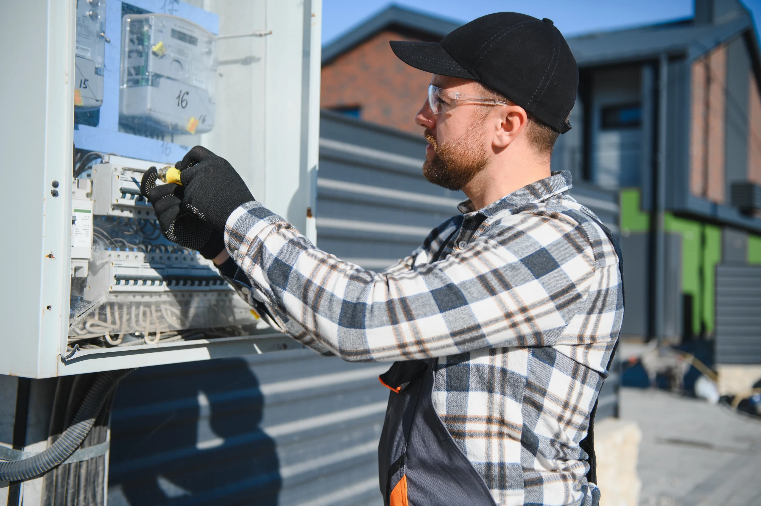 Residential electrician repairing an electrical panel outdoors in a plaid shirt and gloves