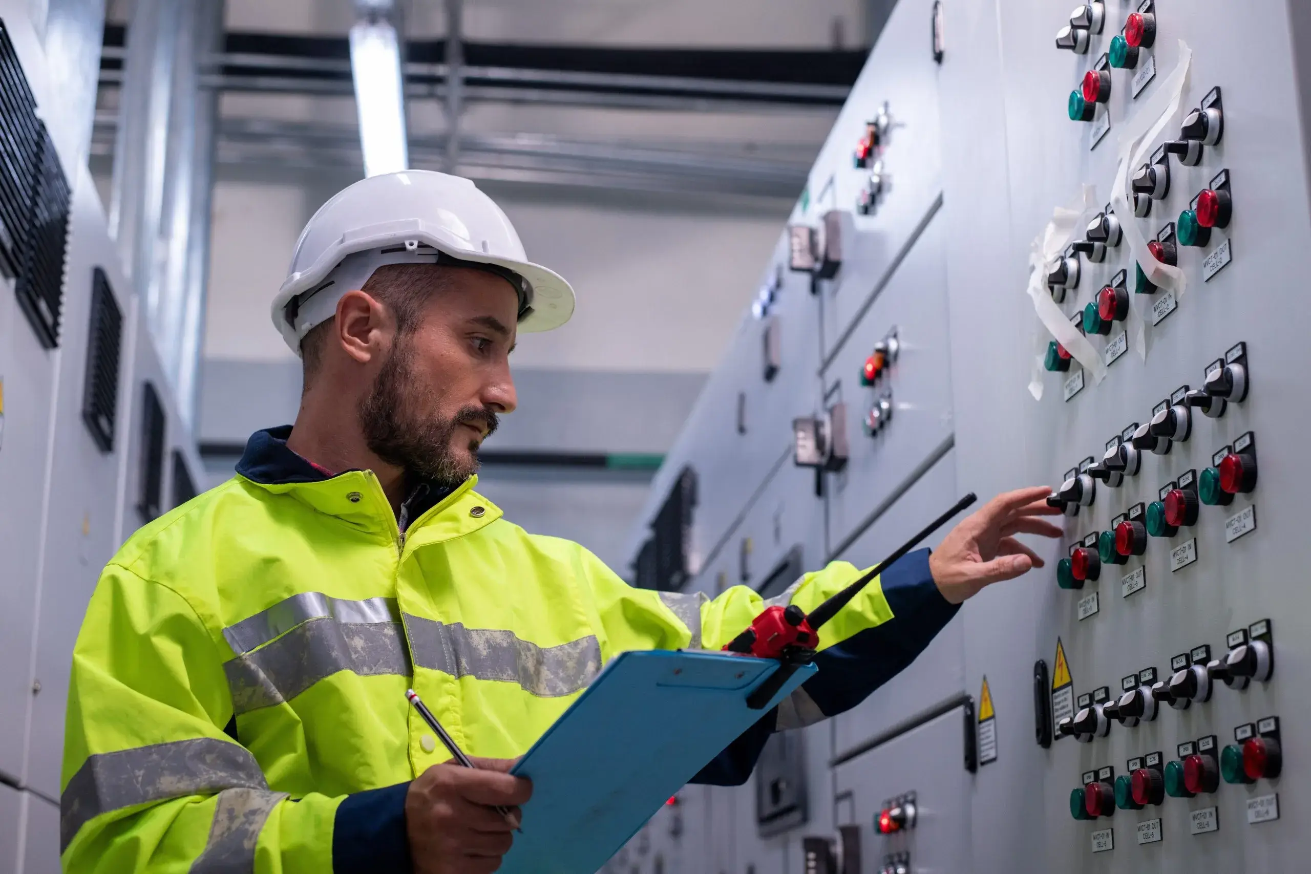 A worker in safety gear examines control panels while holding a clipboard