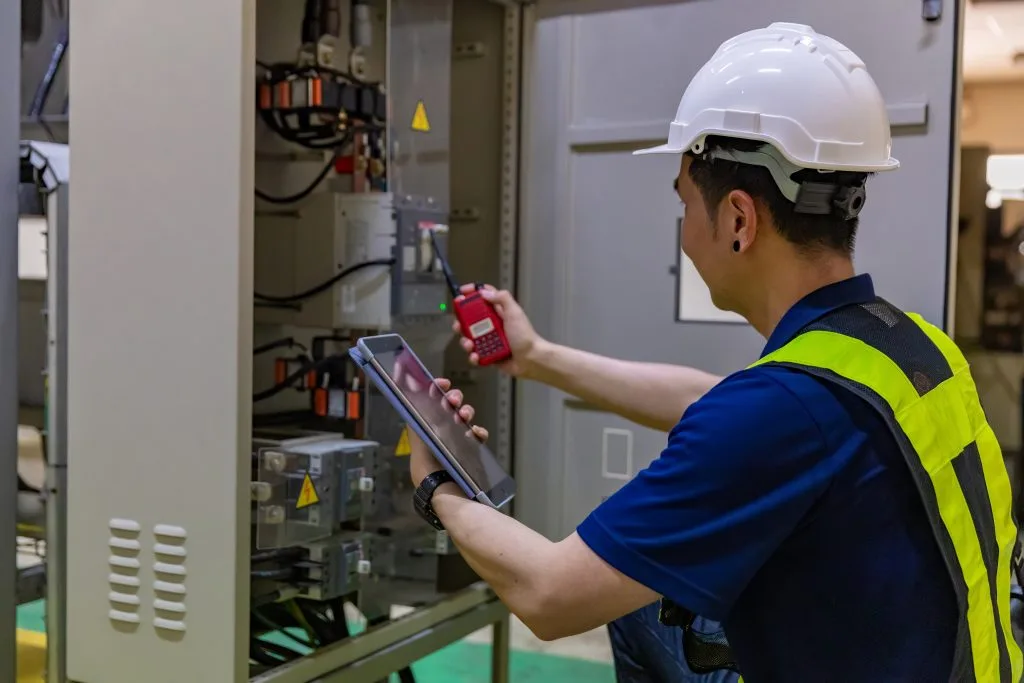 A worker in a hard hat uses a tablet while inspecting electrical equipment